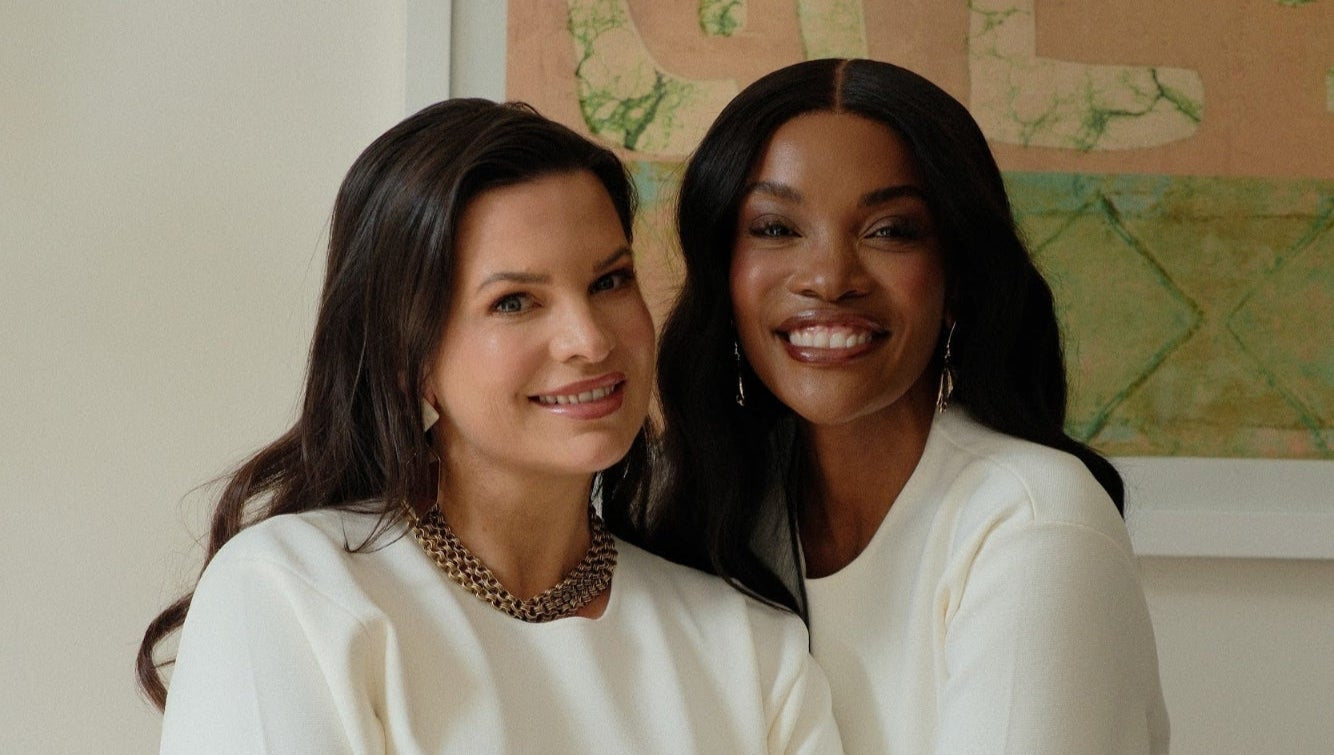 Two women in white dresses sitting together with a neutral background